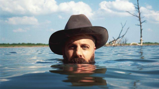 Marty Bush rising up out of a lake while wearing a cowboy hat. He is submerged up to his beard and facing the viewer.